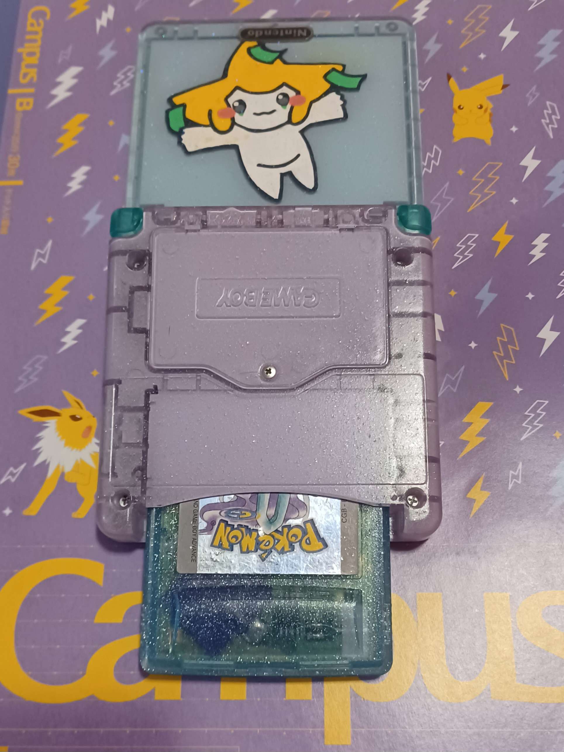a GBA SP, the top half pastel blue with jirachi, and the bottom half pastel purple. the buttons are the same translucent teal as the cartridge in the slot, pokemon crystal.